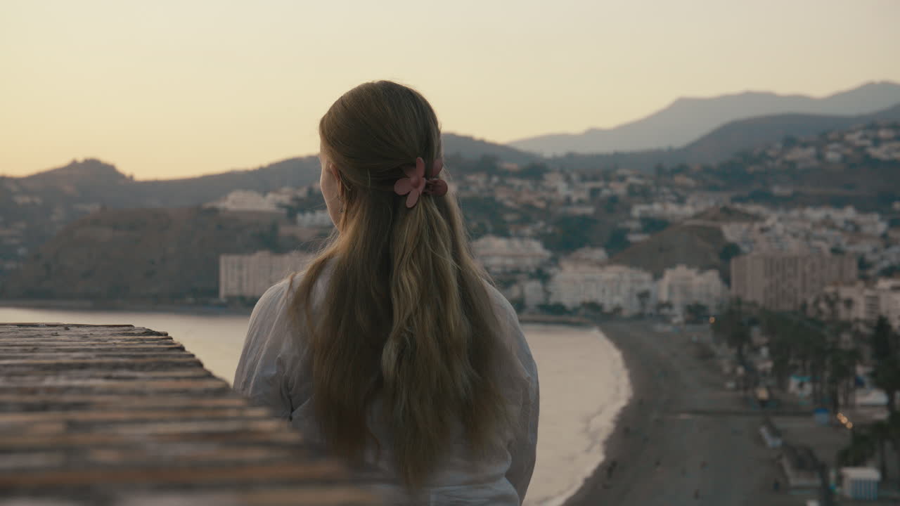 Over-the-shoulder medium shot with shallow focus, capturing a woman watching the city and beach fade into evening. Quiet, thoughtful, and introspective mood