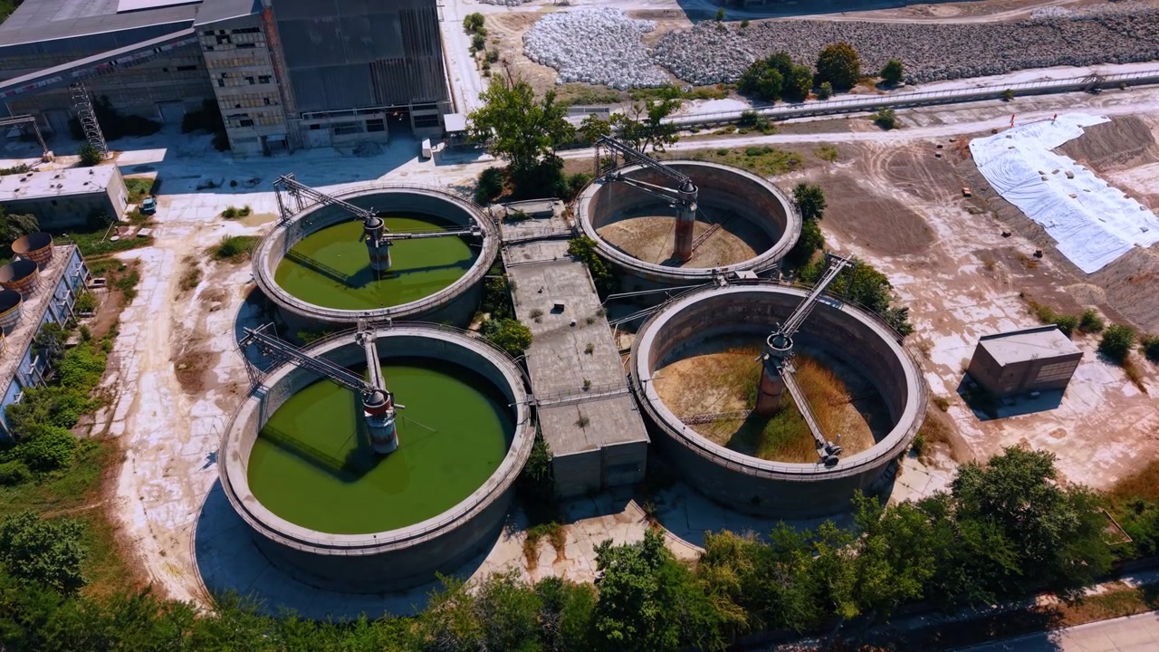 Circular industrial water treatment tanks from an aerial view. Two clarifiers contain green liquid while two remain dry inside a large factory compound on a sunny day.