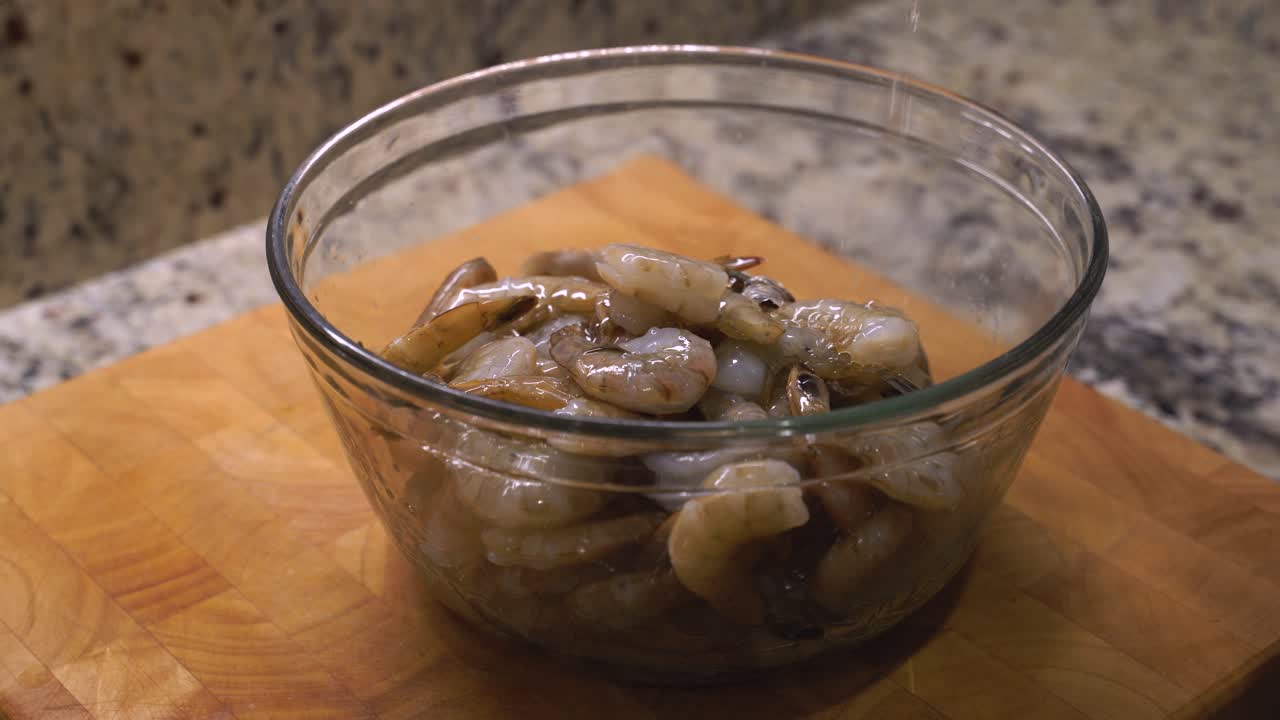Pouring raw unpeeled deveined jumbo shrimp seafood on out of a bag into a large clear bowl, preparing it for cooking and grilling on a wooden butcher block and granite counter top in a kitchen