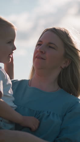 a woman in a blue dress carries her young son, who is wearing a white top, as he smiles brightly. they share a joyful moment outdoors on a sunny day, with soft light illuminating their faces