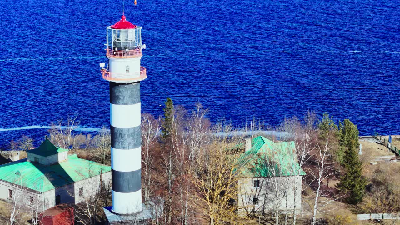 Tall black and white striped lighthouse rises near the vibrant blue sea, surrounded by bare trees and small coastal buildings under clear skies. Ideal for nautical or travel themes