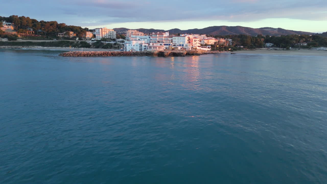 Flying low from the sea to a coastal town atop cliffs. Waves crash against the rocks as the sun reflects on white houses. Stunning aerial view of the coastline, blue waters, and rugged cliffs