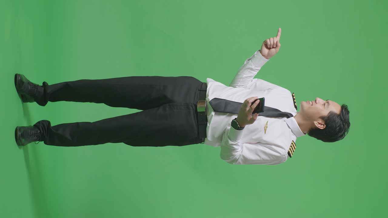 녹색 스크린 배경 스튜디오에 서 있는 동안 스마트폰을 사용하고 을 가리키는 아시아 남자 조종사의 전체 몸