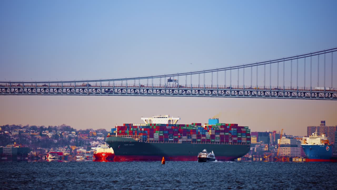 New York, USA, 5 October 2025: Large green container ship sailing under suspension bridge. Cargo vessel passing under a steel bridge at sunset with clear sky and blue water