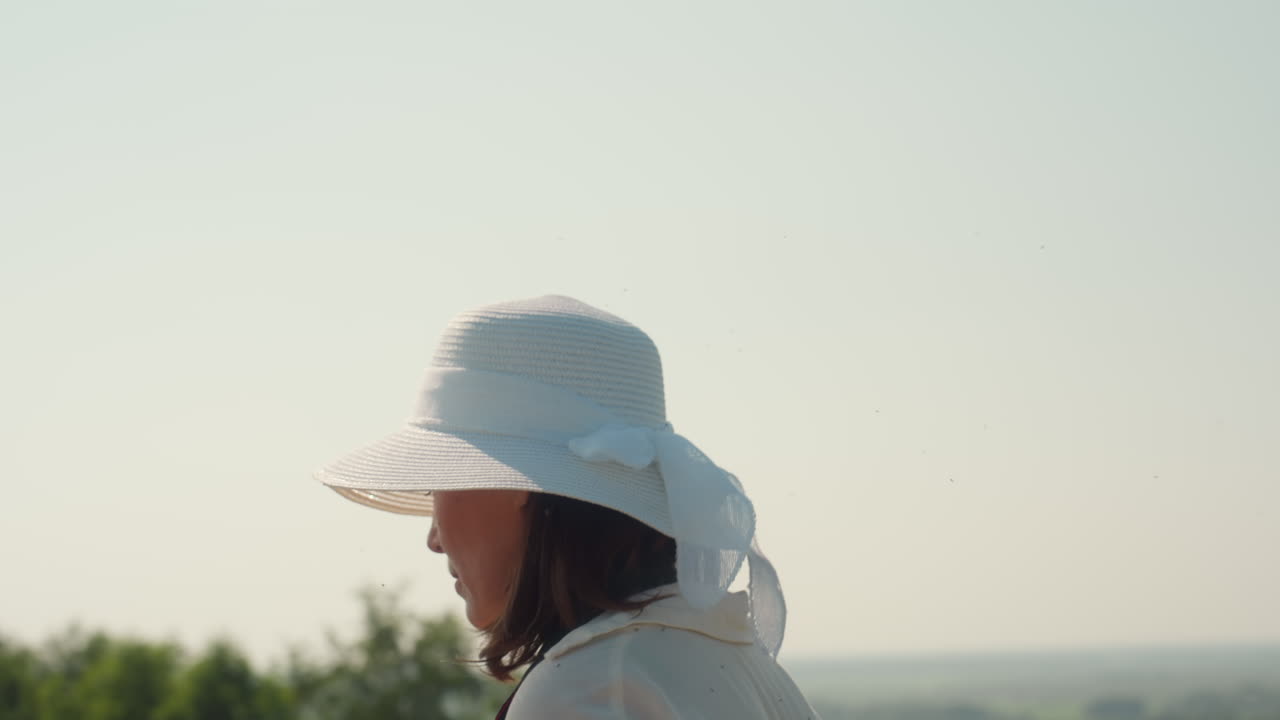 Rear view of woman wearing white sunhat with ribbon walking through sunlit farmland, standing near horizon under pale morning sky, surrounded by treetops and distant greenery