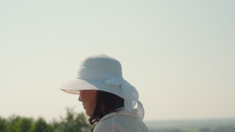 rear view of woman wearing white sunhat with ribbon walking through sunlit farmland, standing near horizon under pale morning sky, surrounded by treetops and distant greenery