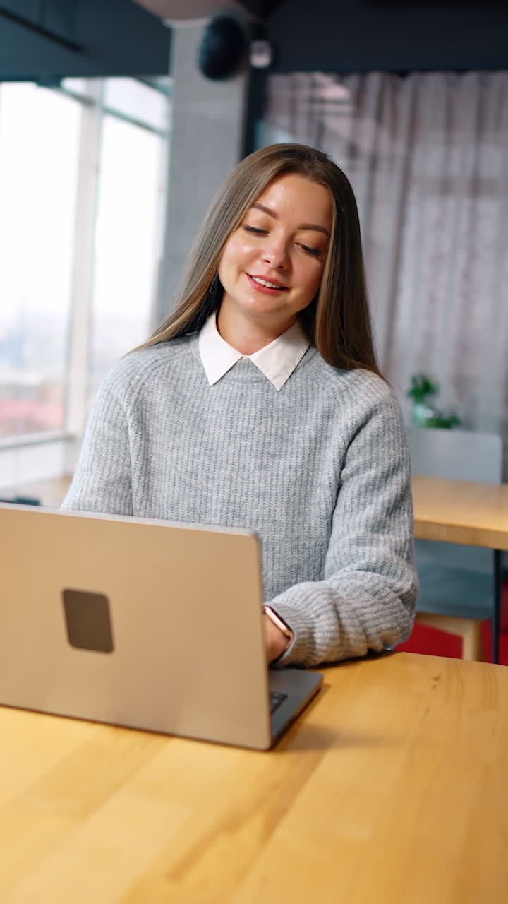 Focused Caucasian woman with long brown hair sits at the desk. Office employee types on her laptop and smiles. Vertical video