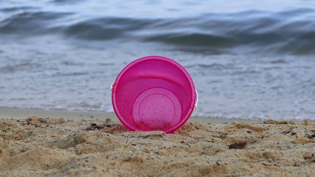 A pink bucket on the sand at the beach with waves hitting the shore on the background