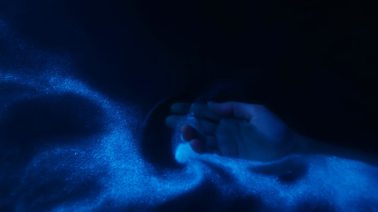 Sabbia blu luminosa che scorre tra le mani, primo piano di polvere fine e particelle scintillanti sotto una luce soffusa.