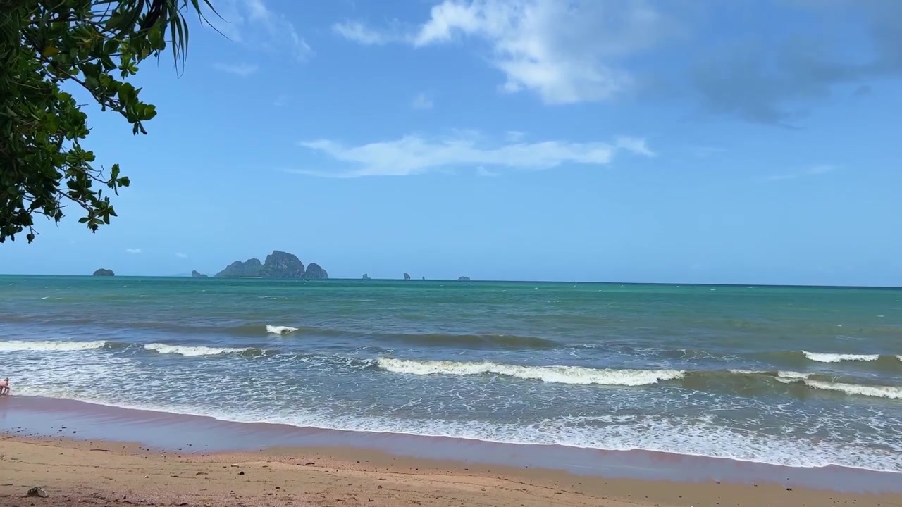 ภาพทิวทัศน์นิ่งของชายหาดเขตร้อนอันเงียบสงบในกระบี่ ประเทศไทย พร้อมคลื่นทะเลที่อ่อนโยนซัดเข้าสู่ชายฝั่งทราย และเกาะหินปูนที่อยู่ไกลออกไปมองเห็นได้ตามแนวขอบฟ้า