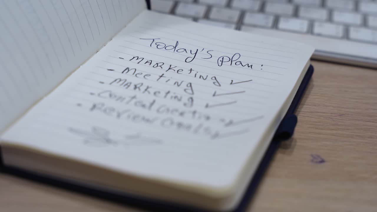 hand closing a notebook with a handwritten daily plan checklist on a wooden office desk, with keyboard in soft focus and shallow depth of field