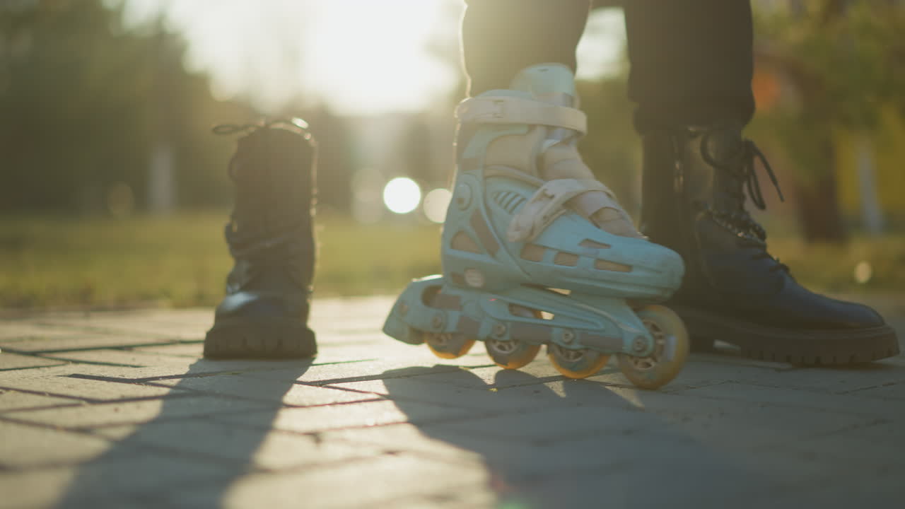 A close-up shot of a person wearing a light blue rollerblade on one foot, with the rollerblade wheels in motion on a paved path in a park during a sunny day. The other foot is in a black boot
