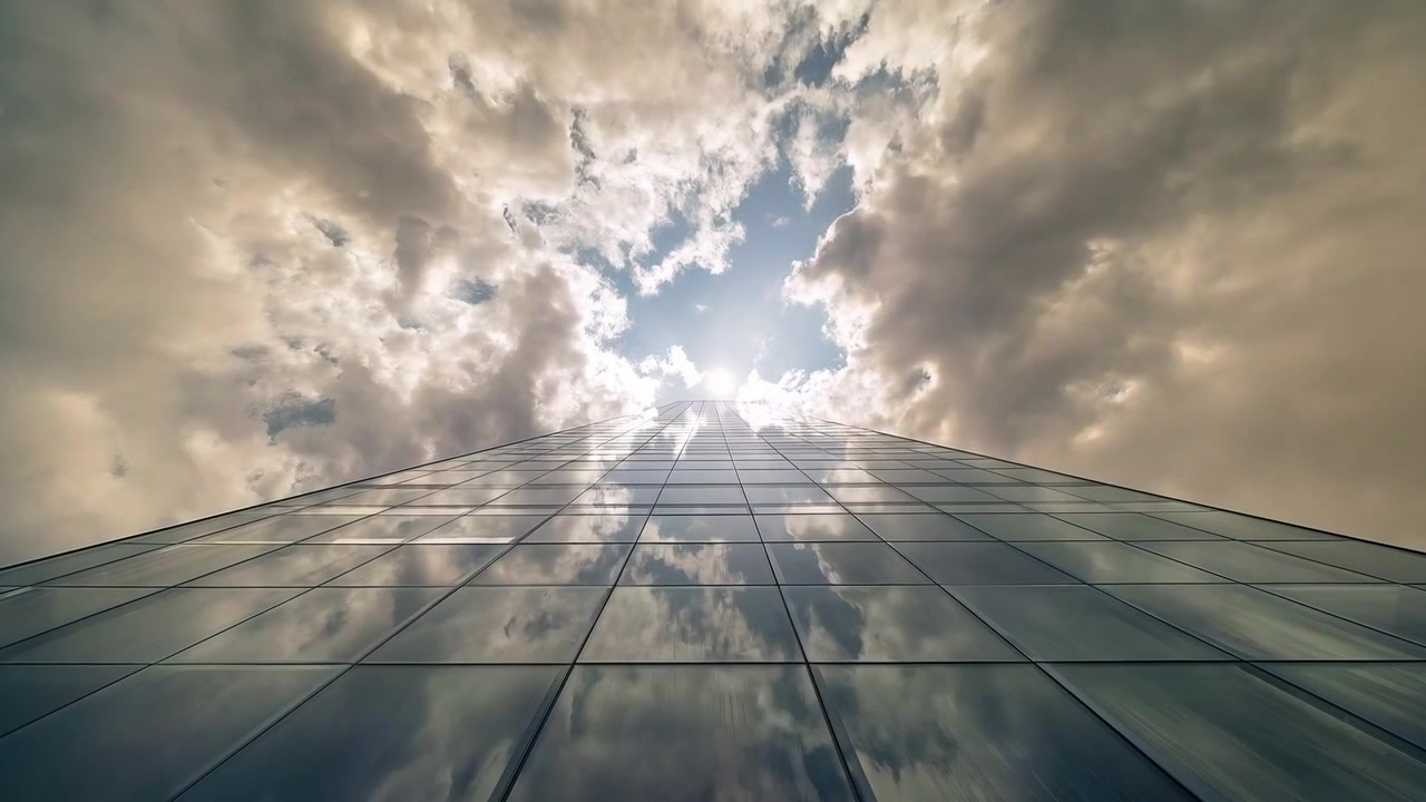 Telecamera in movimento che riprende una facciata a vetri dalla strada, che riflette le nuvole, con il sole che filtra attraverso i montanti.