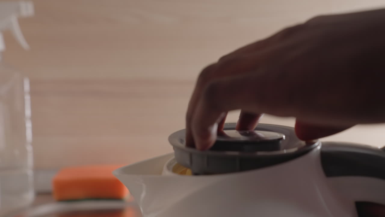 Close-up of man pouring water from electric kettle into sink. Water flows from kettle spout, captured in a moment of morning routine. Cleaning supplies and sponge in background