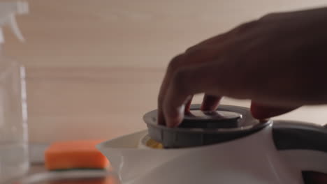 close-up of man pouring water from electric kettle into sink. water flows from kettle spout, captured in a moment of morning routine. cleaning supplies and sponge in background