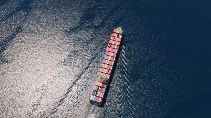 Aerial view from above of a massive container ship transporting cargo across the open ocean, with sunlight reflecting on the deep blue water and creating a beautiful shimmering effect