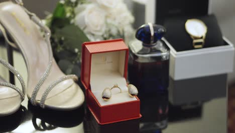 a red box with earrings and a wedding ring next to a perfume bottle and a watch