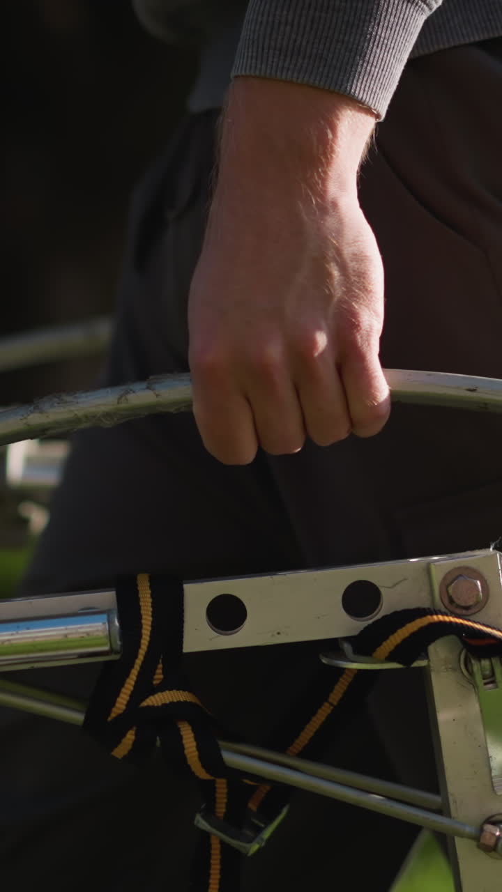 primer plano de una mano humana agarrando una barra de metal, mostrando la mano, la parte inferior del brazo en una camisa gris de manga larga, pantalones de trabajo marrones gris oscuro. el fondo de hierba verde borroso sugiere un entorno al aire libre