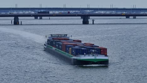 Buque-Portacontenedores-Verde-Y-Blanco-Moviéndose-Sobre-Un-Río-En-Holanda-Con-Puentes-En-El-Fondo