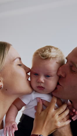 adorable blond blue-eyed baby boy in hands of his parents. loving parents kiss their beloved infant on the cheeks. vertical video