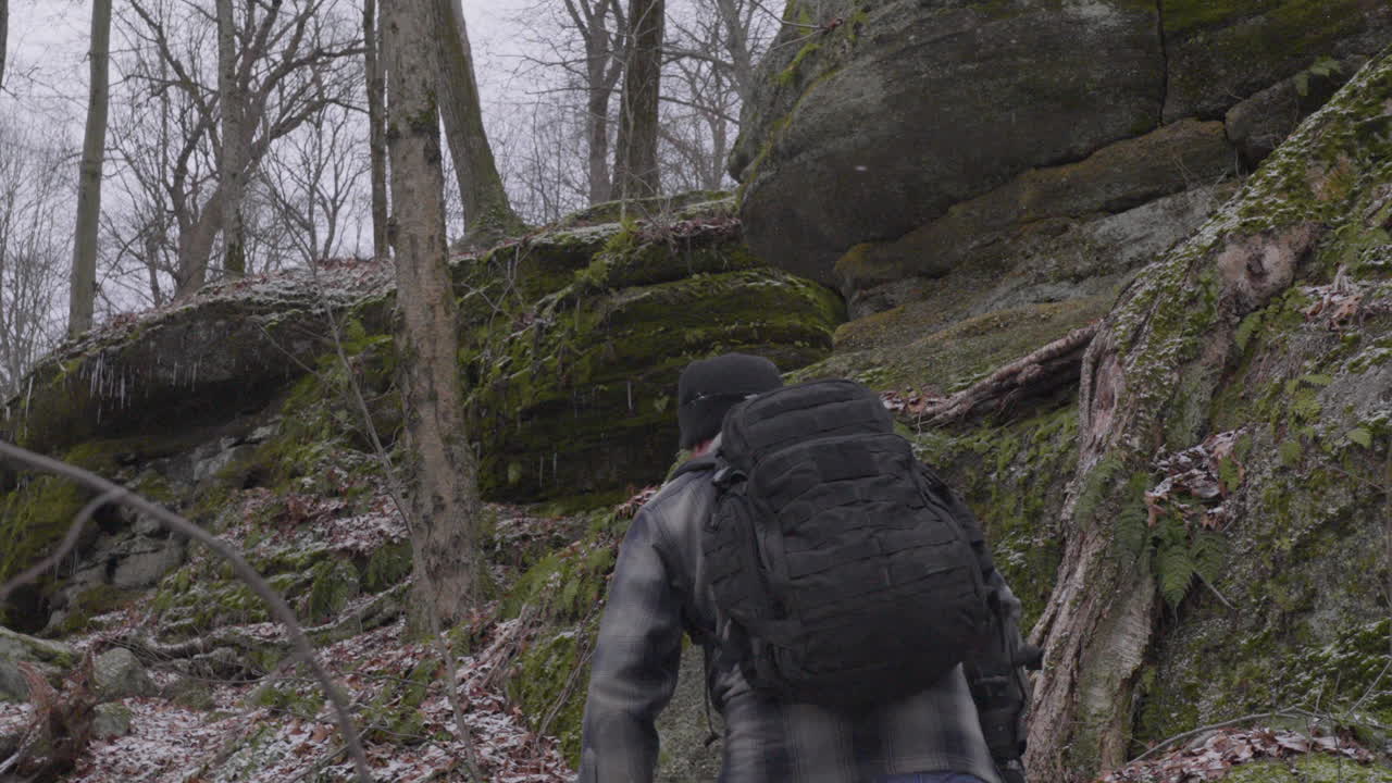 En vandrer med en sort rygsæk og fuldskæg klatrer op ad en stenet bjergside blandt mosbegroede klipper og nøgne træer i Ohio på en grå vinterdag