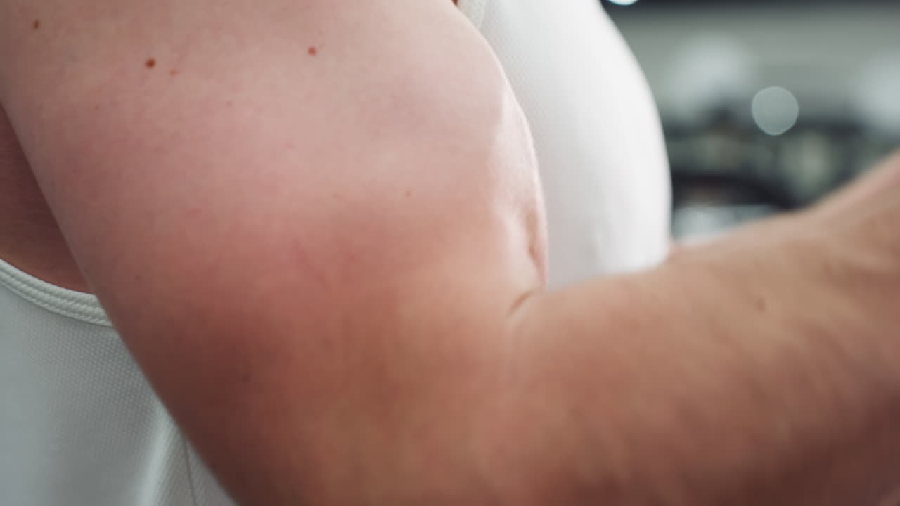 Young man arm and chest muscles flexing under tension on cable machine handle during intense gym workout, white singlet clinging to skin, showcasing power and form in modern fitness environment