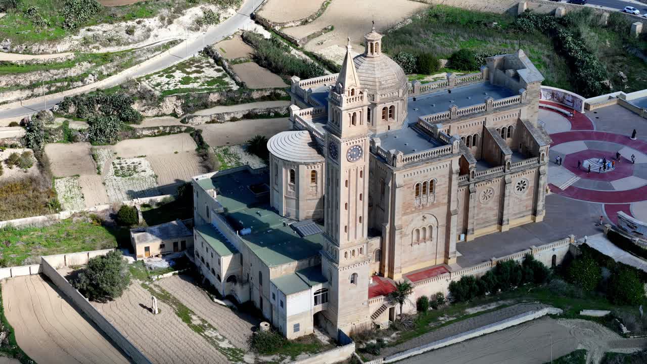 Açık güneşli bir yaz gökyüzünün altında, uzun çan kulesi ve dairesel mozaik meydanıyla ikonik Romanesk mimariyi sergileyen Gozo Malta'daki görkemli Ta Pinu Tapınağı'nın havadan yüksek açılı görünümü.