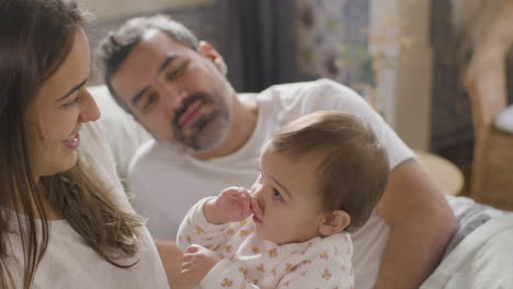 Mujer-Feliz-Jugando-Con-Su-Hija-En-La-Cama-Mientras-Su-Esposo-Los-Observa