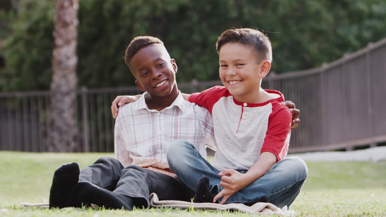 Due amici maschi preadolescenti seduti sull'erba, abbracciati e sorridenti alla macchina fotografica.