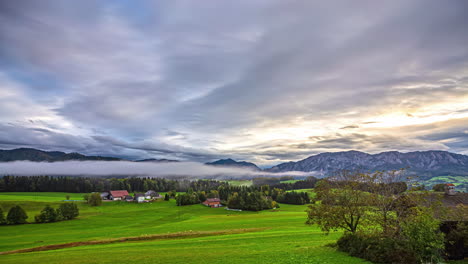 Zeitraffer,-Wolken-Ziehen-Schnell-über-Grüne,-Nasse-Landwirtschaftliche-Wiesen,-Die-Sonne-Scheint-über-Bäumen-Und-Lichtstrahlen-Durch-Die-Neblige-Skyline