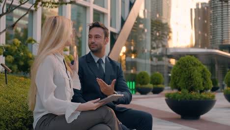 two business colleagues man in 30s and woman in 20s having conversation in park outside corporate building entrance sitting on bench and using tablet - medium shot, slow motion