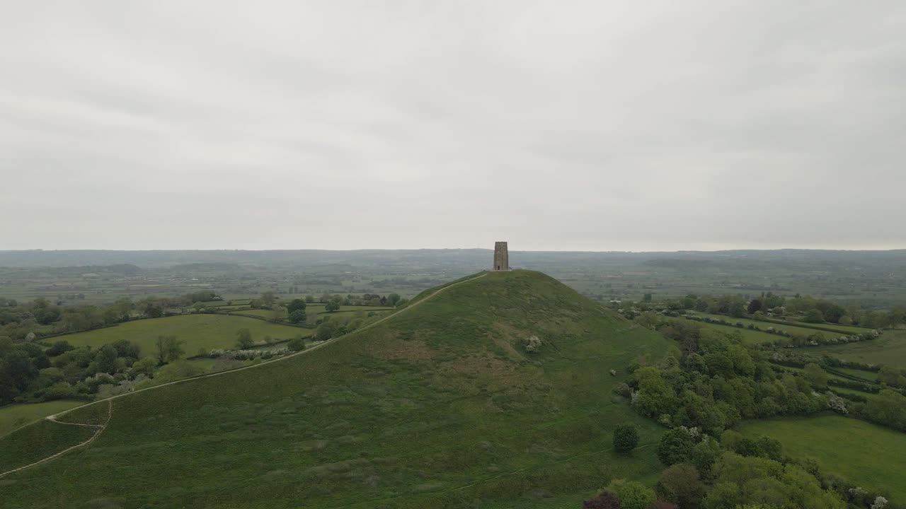 Aerial view of Glastonbury, drone rotating and camera facing down over the Glastonbury Tor, drone flying over the green fields with some houses on the left side of the camera. 4K, 60fps.