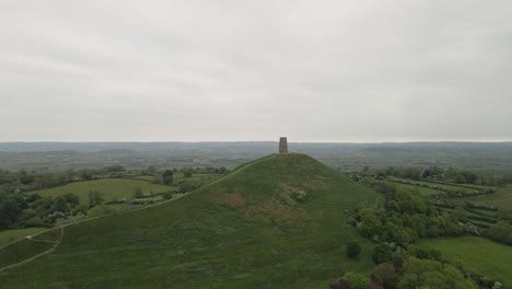 aerial view of glastonbury, drone rotating and camera facing down over the glastonbury tor, drone flying over the green fields with some houses on the left side of the camera. 4k, 60fps.