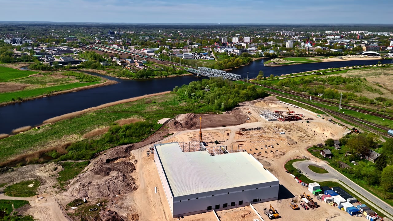 Aerial view of a construction site by a river and railway near the city. Urban development scene with industrial building, bridge crossing and expanding infrastructure under daylight.