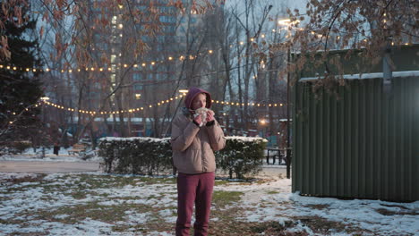 young woman standing outdoors with napkin in hand eating corn, urban park background with snow on ground, holiday lights glowing, casual winter food enjoyment, food break in park