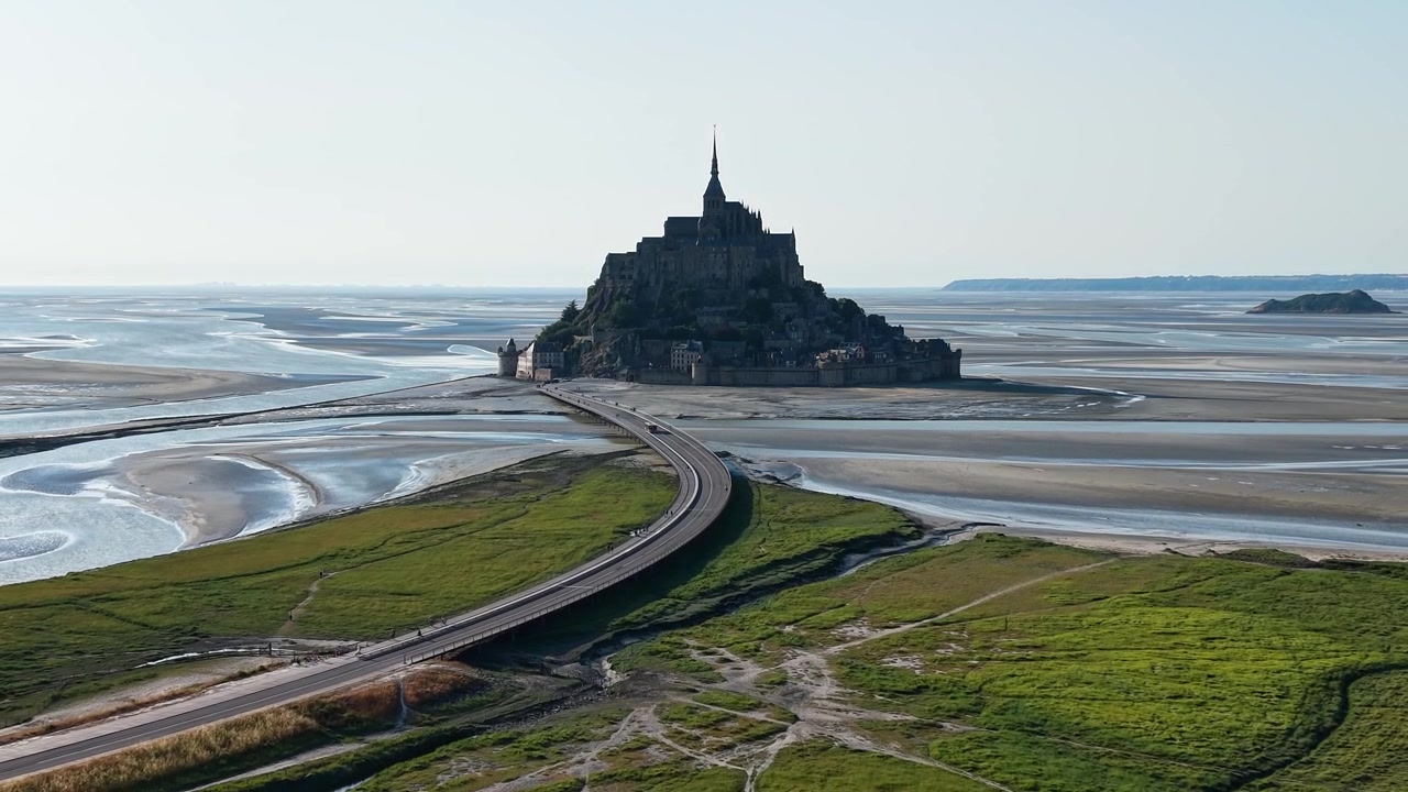 Filmisk dronebillede af Mont Saint-Michel med det ikoniske middelalderkloster, den hævede dæmning, der forbinder øen med fastlandet i Normandiet, og de store vadeflader, der blotlægges ved lavvande.