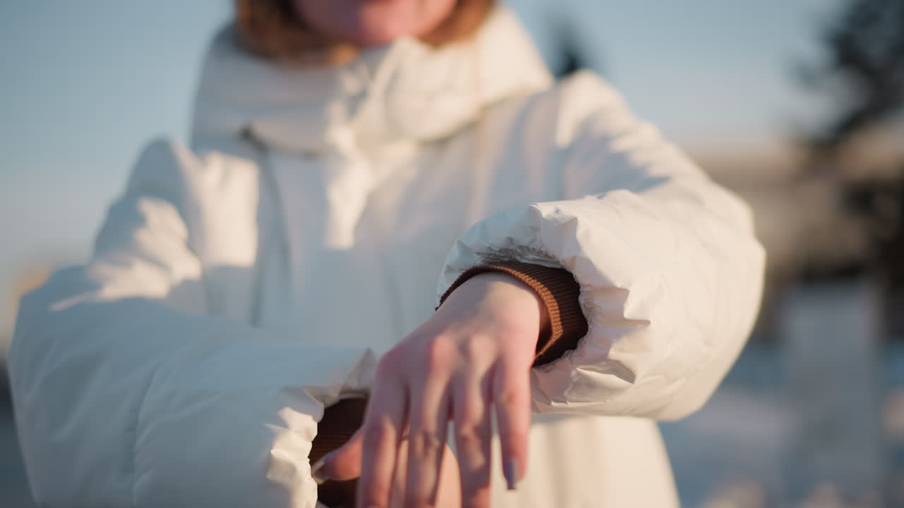 Woman wearing winter coat and beanie expressing herself through hand motion under soft sunset light against blurred forest backdrop with snowy ground and headphones evoking graceful movement and calm
