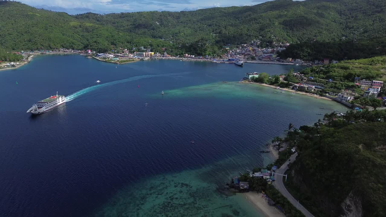 Drone aereo che riprende una grande nave passeggeri in uscita dal porto di Romblon, circondata da acque cristalline color turchese, colline verdi e cielo blu, mostrando un paesaggio costiero e isolano tropicale e suggestivo