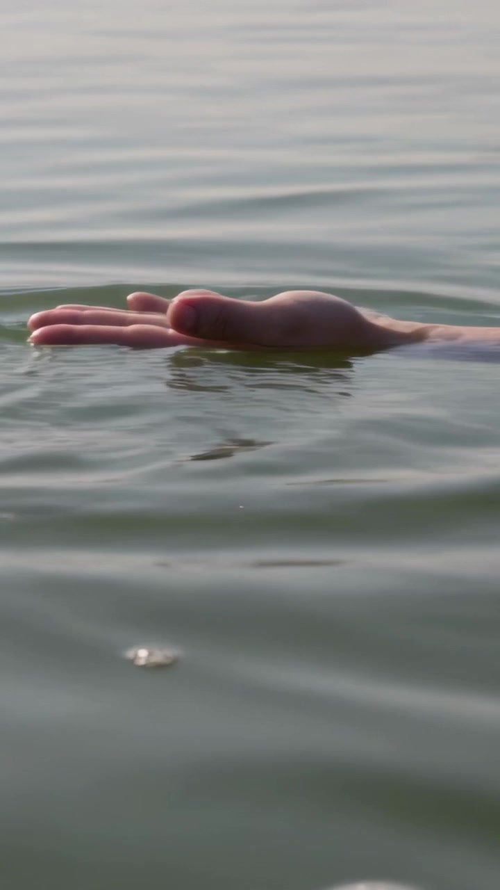 Vertical video: Drifting forearm and hand rising, opening on calm sea as ripples lifting palm