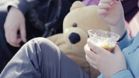 close up of little girl with blue jumper nail polish and teddy bear beside her holding a sundae ice cream sitting outdoor 4k