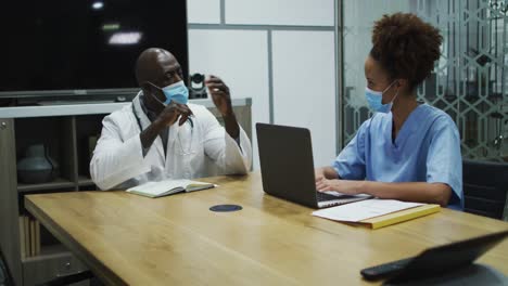 Diverse-female-and-male-doctors-wearing-face-masks-discussing-in-office-using-laptop