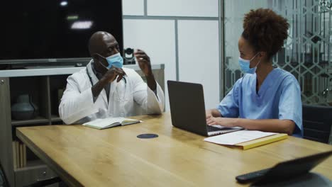 diverse female and male doctors wearing face masks discussing in office using laptop