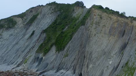 sheer rocky cliffside edges of zumaia spain beach, covered in grassy green shrub, aerial pullback