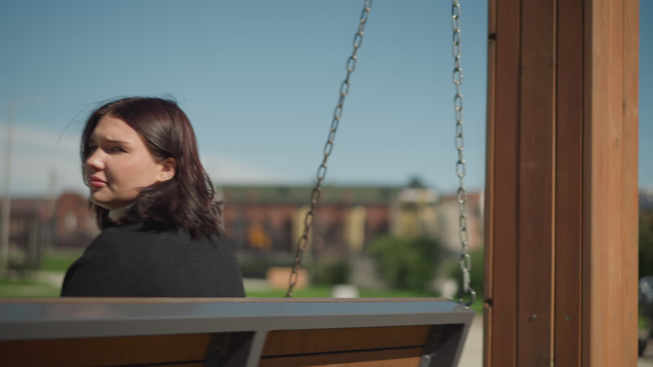 야외에서 나무 스에 앉아있는 여자의 가까운 모습, 그녀의 머리카락은 바람과 함께 약간 움직이며, 밝은 은 날 배경에 건물과 초록색의 흐릿한 시야가 있습니다.