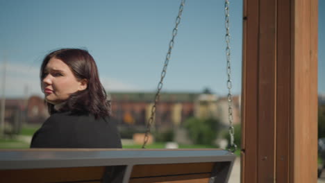 close up de la dama sentada en un columpio de madera al aire libre, su cabello se mueve ligeramente con la brisa, con una vista borrosa de un edificio y vegetación en el fondo en un día soleado brillante