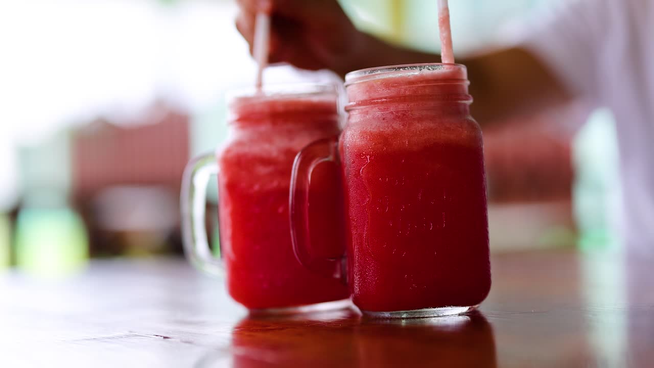 Hands serve vibrant watermelon juice in glass mugs, capturing a refreshing moment in a bright, casual setting