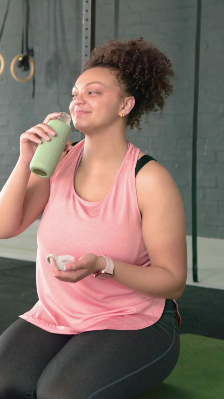 Video verticale: Donna in top rosa inginocchiata su un tappetino da palestra, controlla il tappo e si idrata sorseggiando da una bottiglia verde.