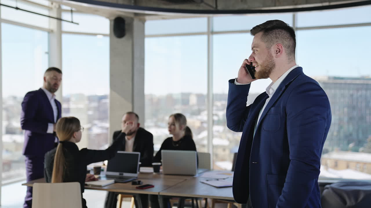 Serious businessman having conversation over the phone. Coworkers' workplace in office at the background.
