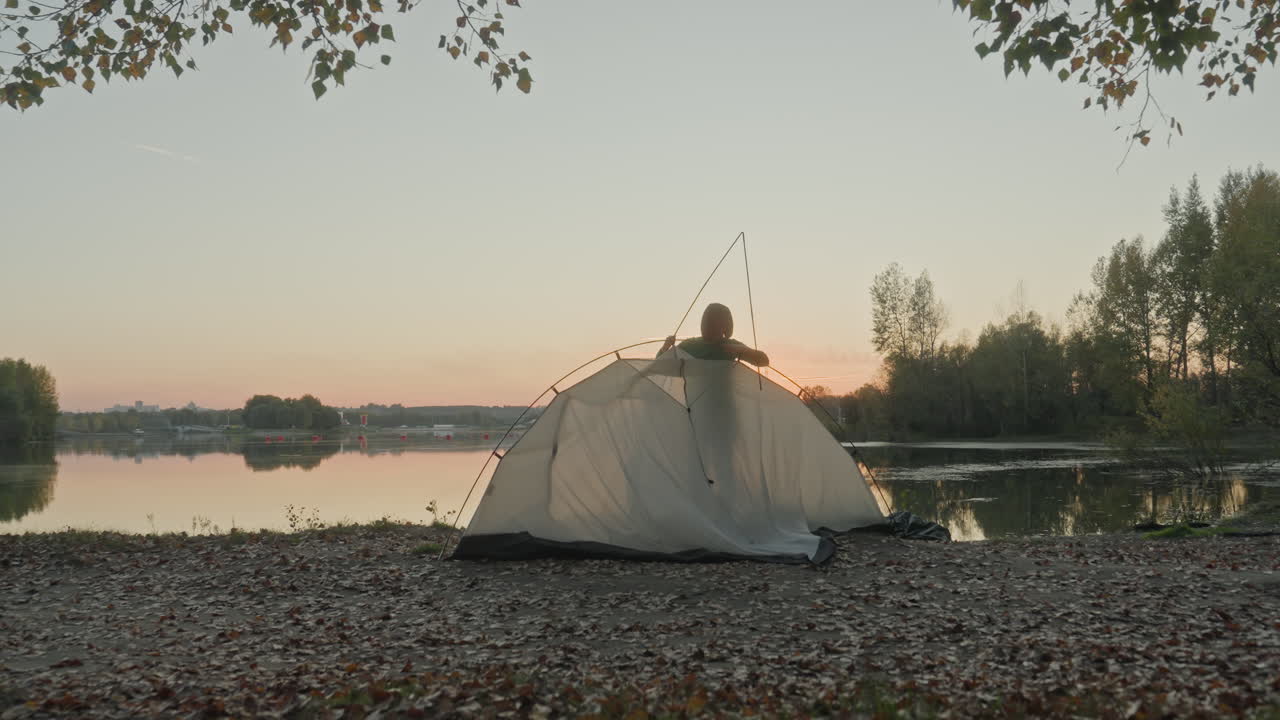 Fredfyldt lejr ved søen under solnedgangshimmel. Ensom campingplads langs fredelig stenet søbred ved skumringstid. Afsidesliggende telt ved søen placeret blandt klipper med roligt vand og pastelfarvet himmel om aftenen