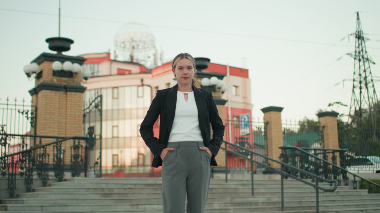 Young lady in professional attire stands confidently with hands on waist in urban setting, posing on steps in front of ornate building with metal railings and brick pillars during bright clear day